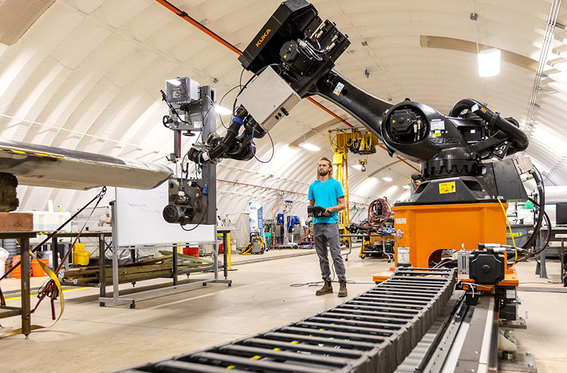 NREL robotics engineer and lead author of the study, Hunter Huth, stands beside the robot programmed to automate part of the wind turbine manufacturing process